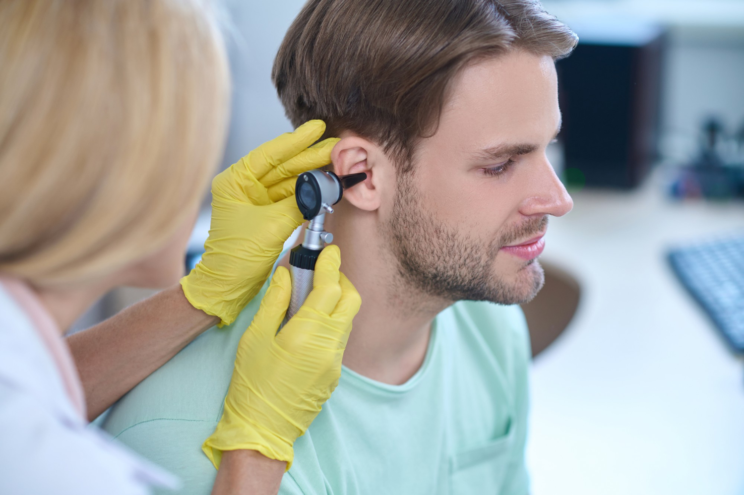 53026 A doctor in yellow gloves examines a man's ear with an otoscope in a medical office.