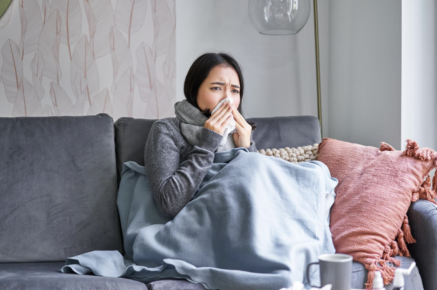 Woman sitting on sofa under a blanket, blowing her nose with a tissue, appearing to have a cold.